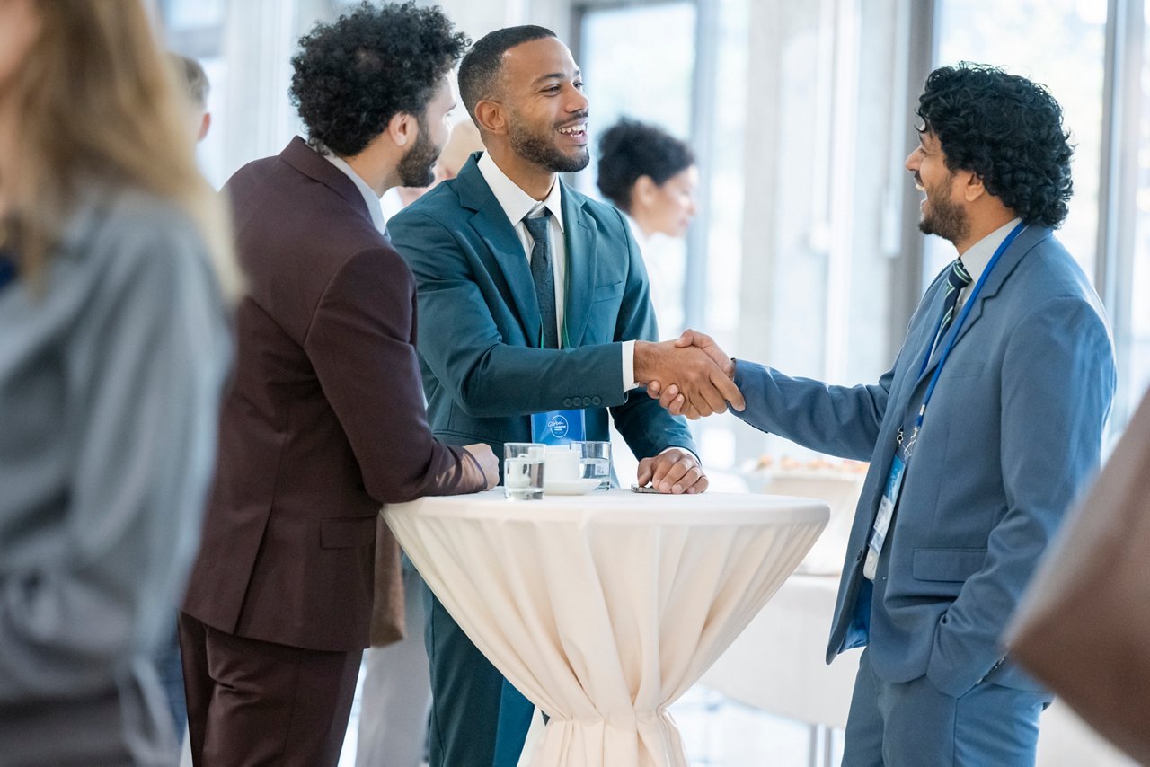Multiracial businessman engaging in a handshake with potential partners at a business conference. The setting includes a networking table with water glasses and the atmosphere is professional and collaborative.