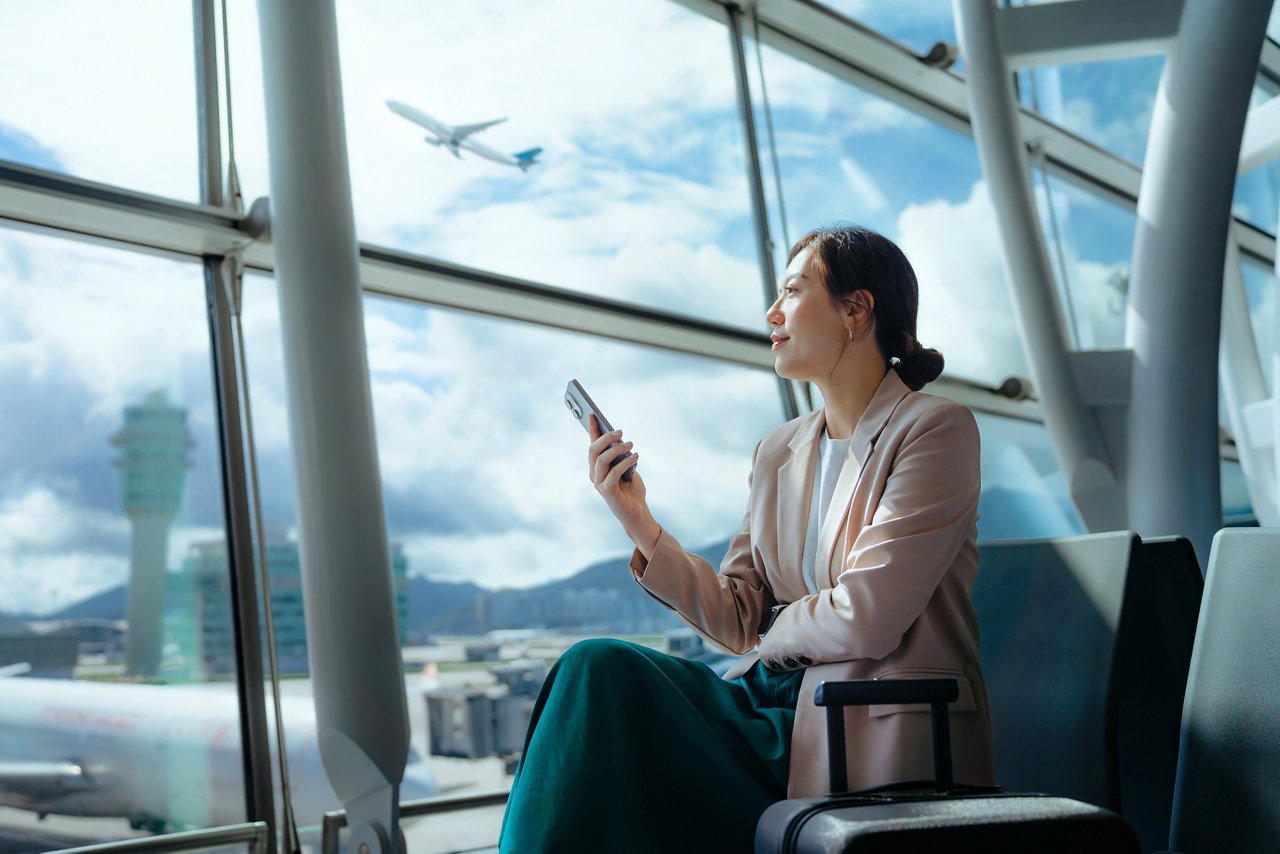 Confident young Asian businesswoman with suitcase looking ahead while using smartphone waiting for her flight at airport terminal. Business traveller. Woman in business. Business lifestyle and technology. Travel and vacation concept