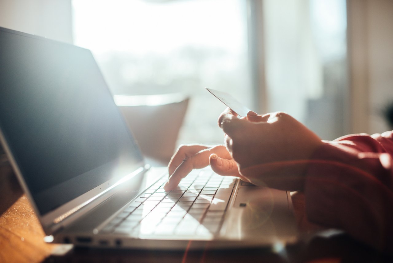 A close-up shows a hand typing on a laptop keyboard while another hand holds a credit card in bright window light
