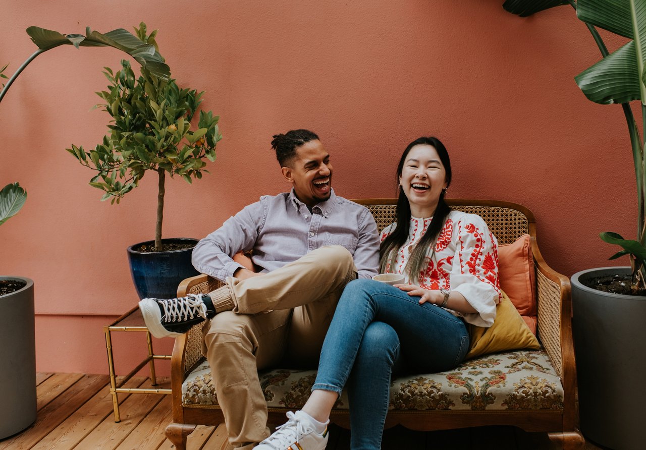 A black man and a Chinese woman sit side by side on a rattan sofa. They are comfortable together as they laugh. Space for copy.