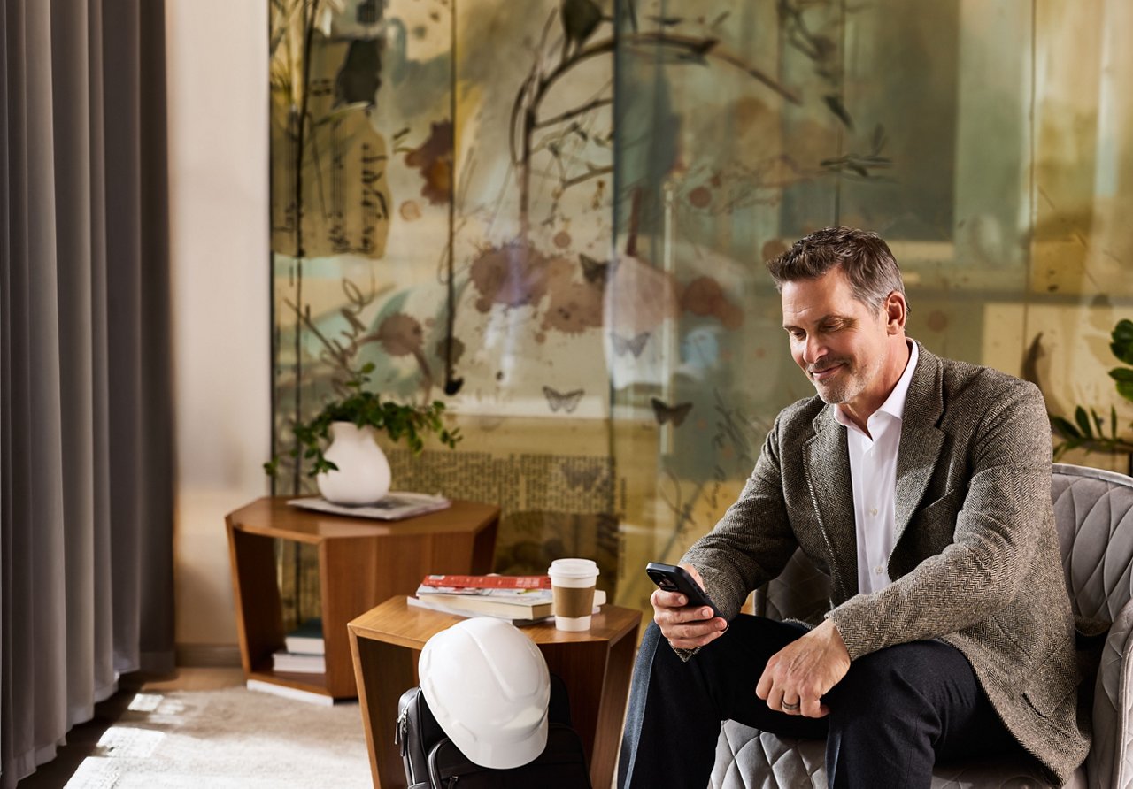 A man in a blazer checks his phone in a lounge chair, a coffee cup and a hard hat resting beside him