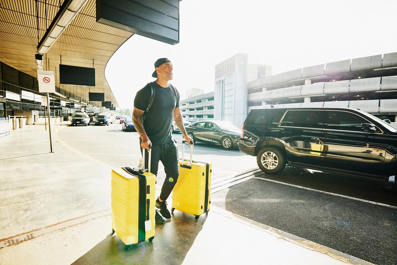 Wide shot of man walking luggage to curb for rideshare pickup at airport after flight