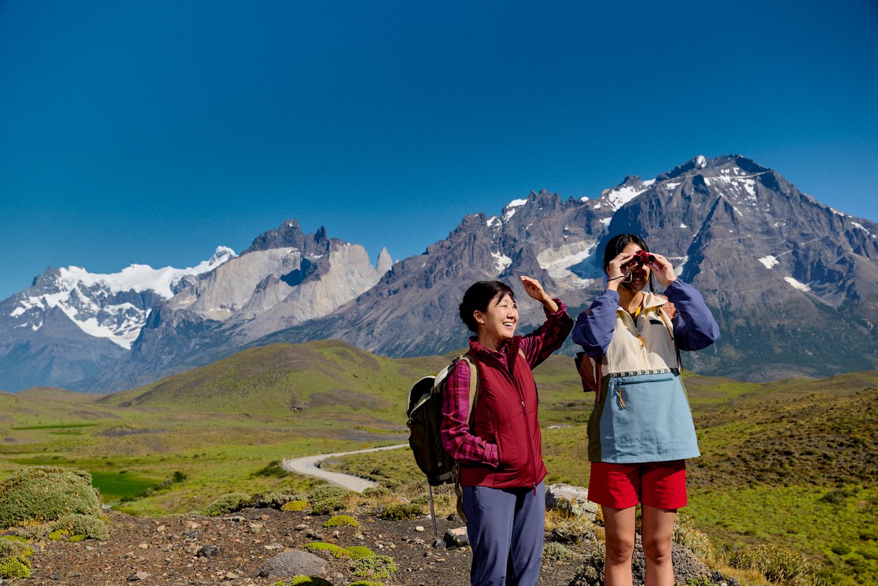 Two hikers with backpacks use binoculars to take in a view of dramatic snow-capped peaks