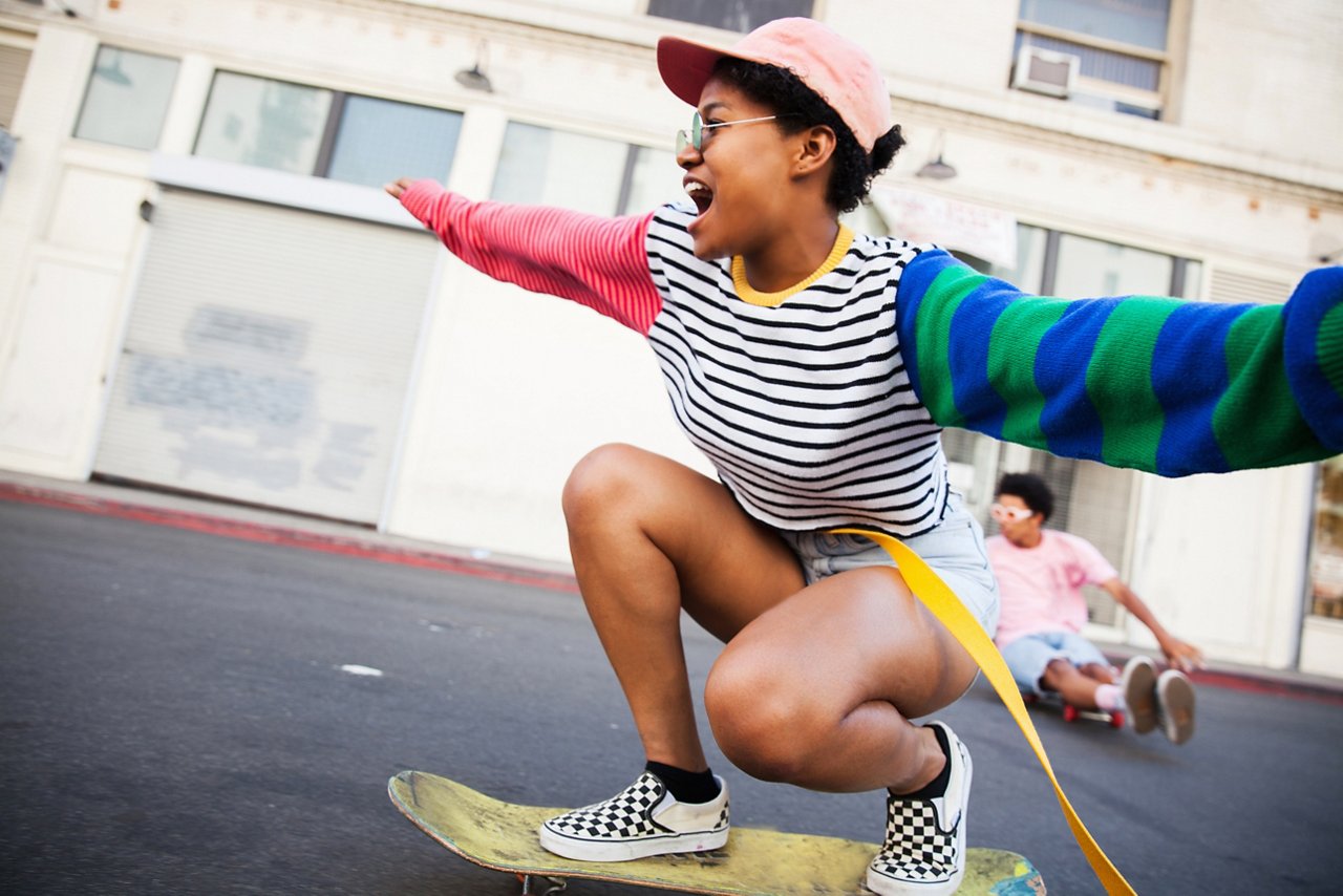 An excited young woman wearing a colorful outfit riding a skateboard down a city street